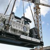 Motor generador industrial de gran escala sin caseta acústica siendo levantado por una grúa torre durante construcción.